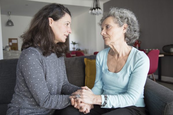 Serious elderly woman and her daughter talking and holding hands Serious elderly woman and her daughter talking and holding hands. Mother and daughter sitting on couch with home interior in background. Daughter and elderly mother concept.